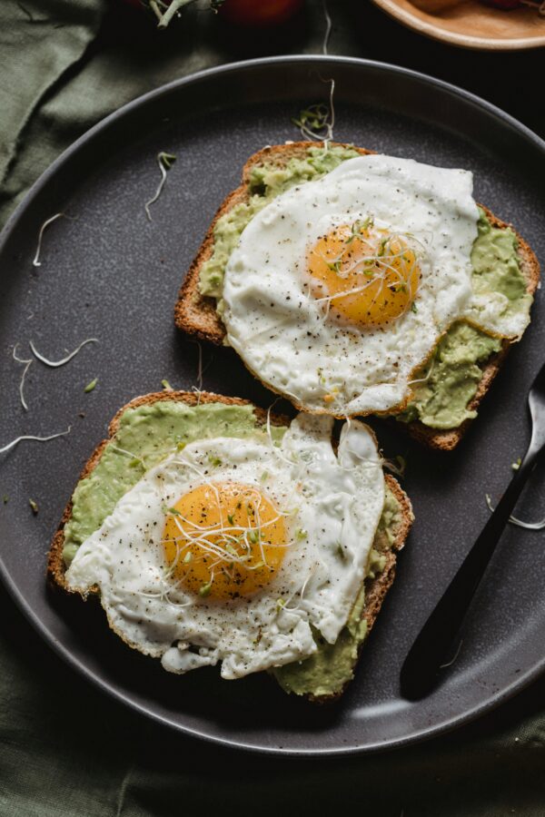 Appetizing avocado toast topped with perfectly fried eggs and sprouts on a dark plate.